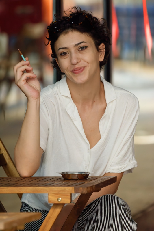 4 - A girl with short, dark curly hair and sunglasses resting on their head, wearing a white button-down shirt and striped pants while sitting at an outdoor wooden table with a small ashtray